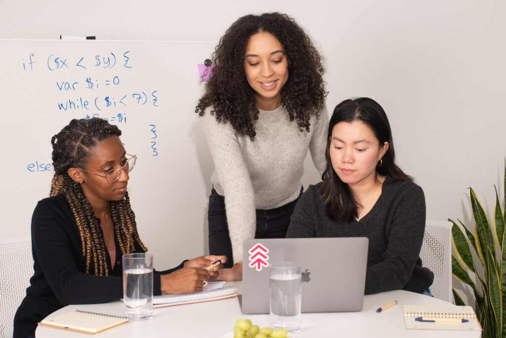 Three people working together, looking at a laptop with a whiteboard in the background.