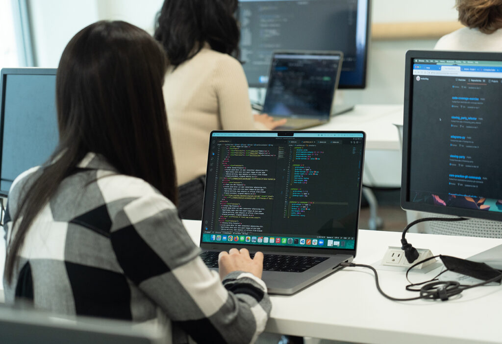 An over-the-shoulder shot of a person with long black hair working on code on a laptop. There are other people working on computers in the background.
